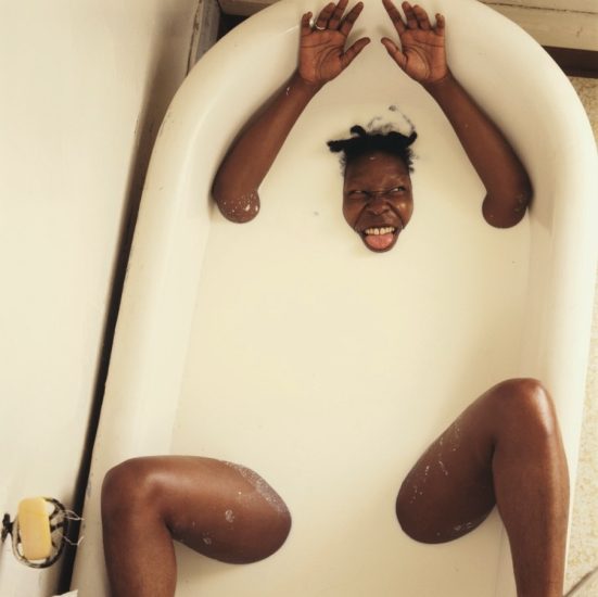 Person submerged in milky bathwater with head, arms, and legs above the surface, grinning playfully and sticking out tongue.